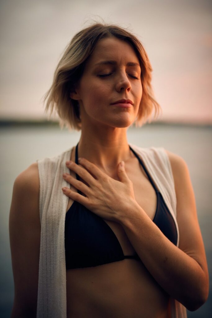 Woman closes her eyes, resting a hand on her chest by the water.