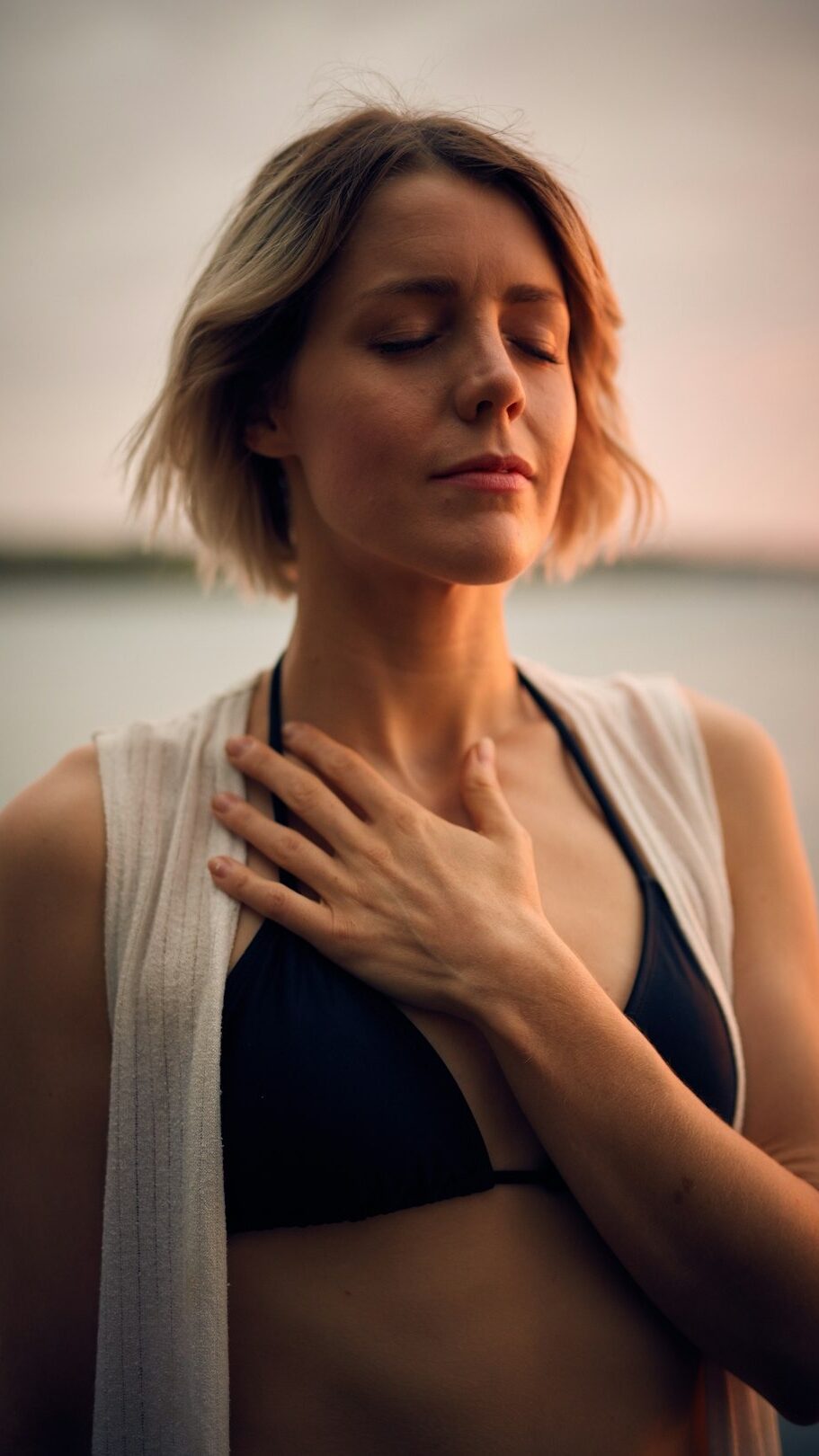 Woman closes her eyes, resting a hand on her chest by the water.